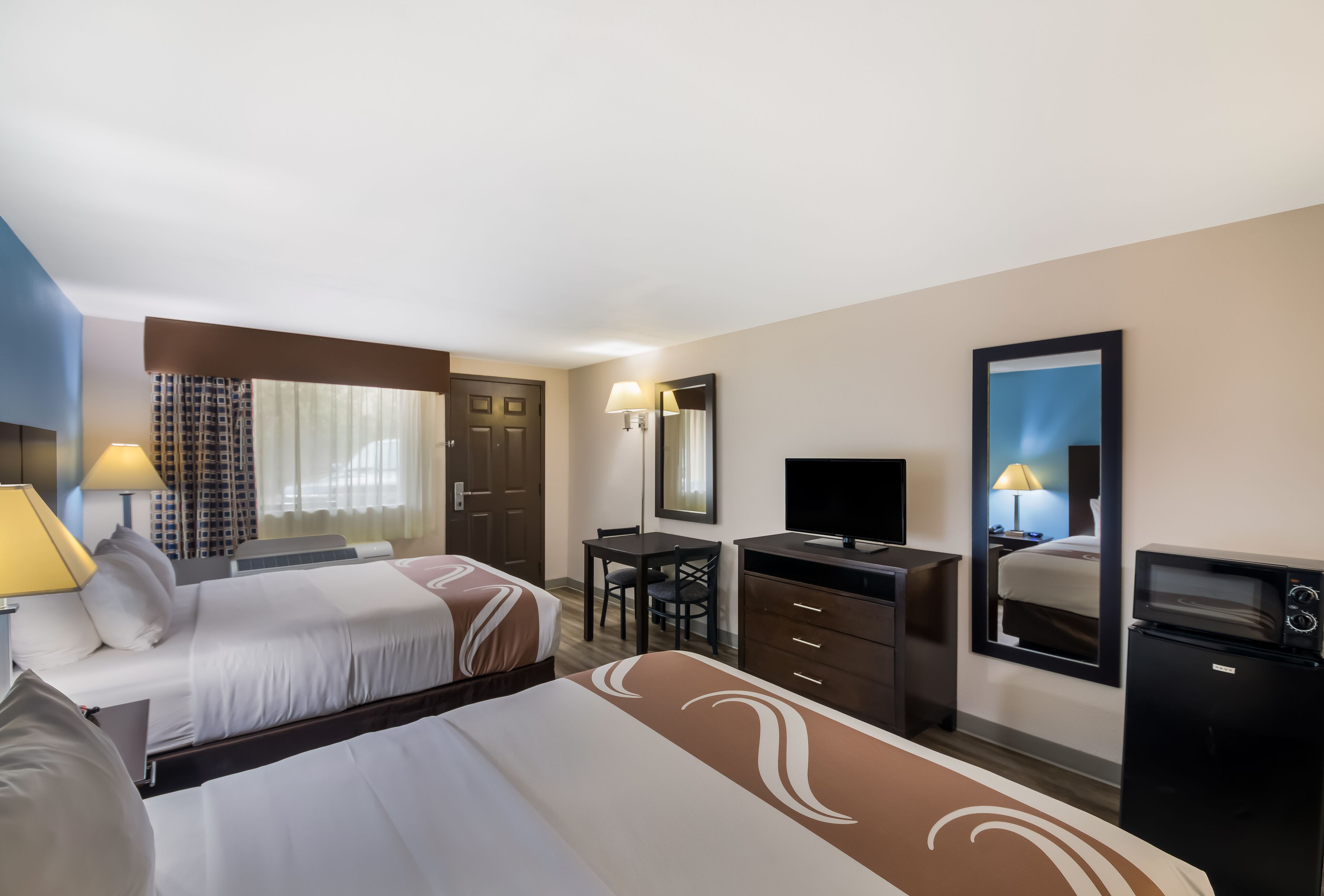 A hotel room with two beds, a desk, a television, a mirror, and a refrigerator.