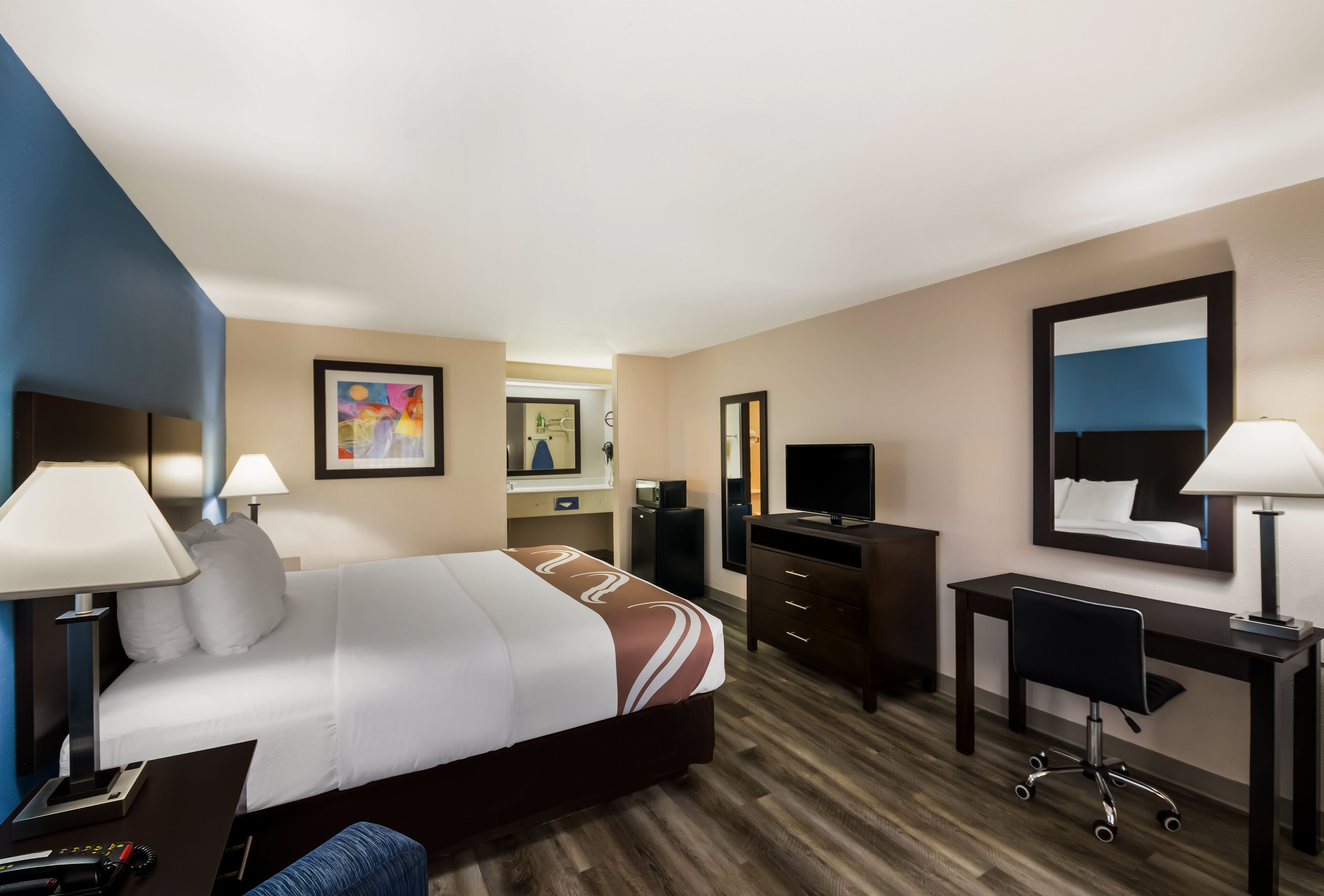 A hotel room with a bed, desk, chair, lamps, and a television.