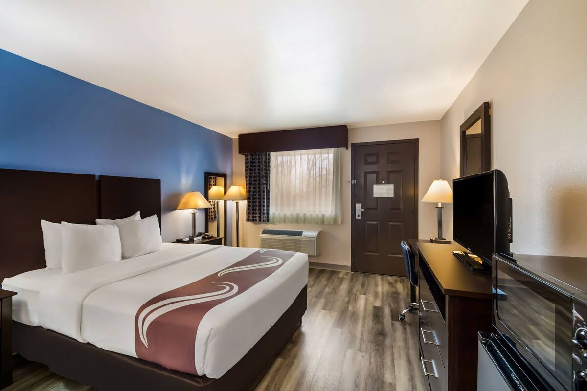 A hotel room with a bed, two bedside lamps, a television, a desk with a computer, and a door.