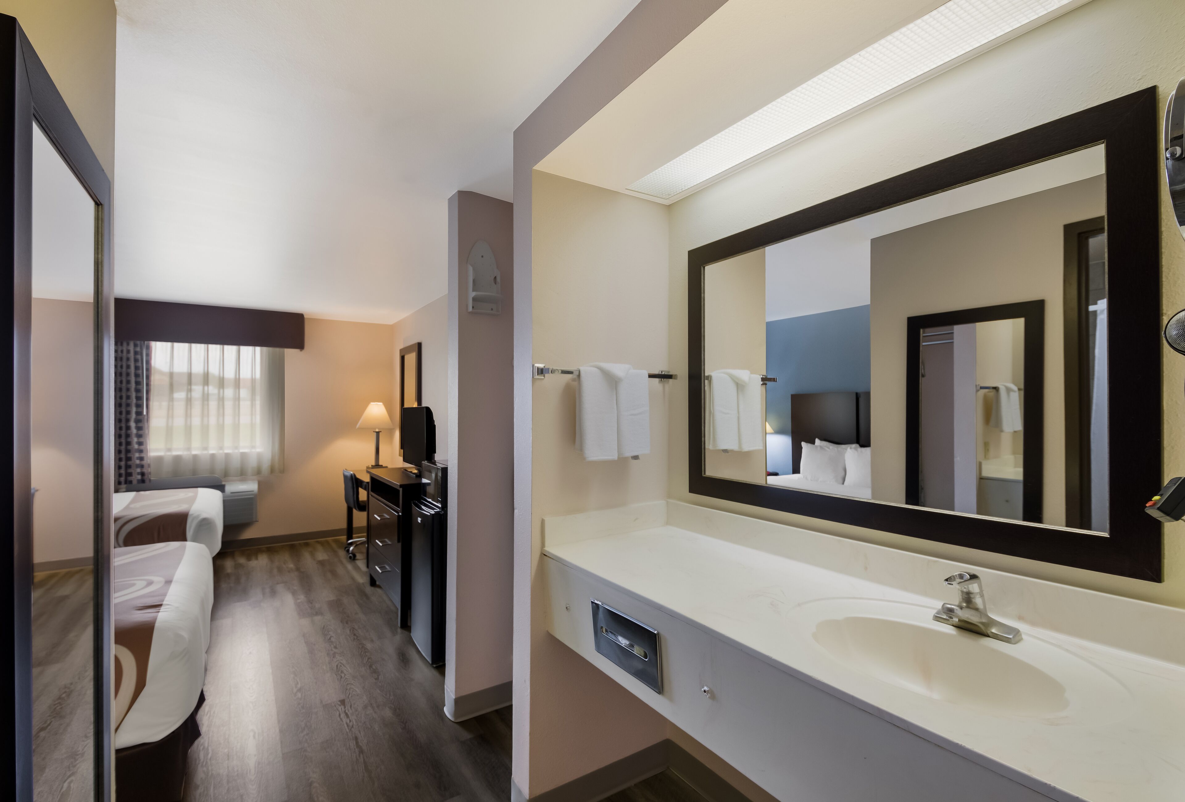 Une chambre d’hôtel avec un grand miroir, un lavabo blanc, un bureau avec une lampe, un lit et une fenêtre avec des rideaux.