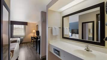 Une chambre d’hôtel avec un grand miroir, un lavabo blanc, un bureau avec une lampe, un lit et une fenêtre avec des rideaux.