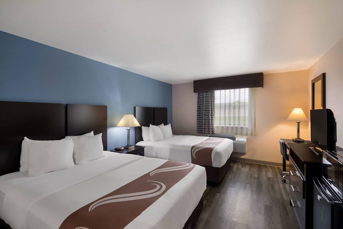 A hotel room with two beds, a desk, a TV, and a window with curtains.