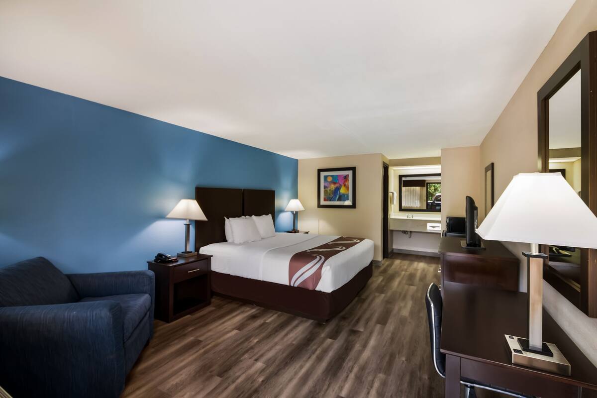 A hotel room with a bed, bedside tables, a desk, a chair, a couch, and a bathroom visible in the background.