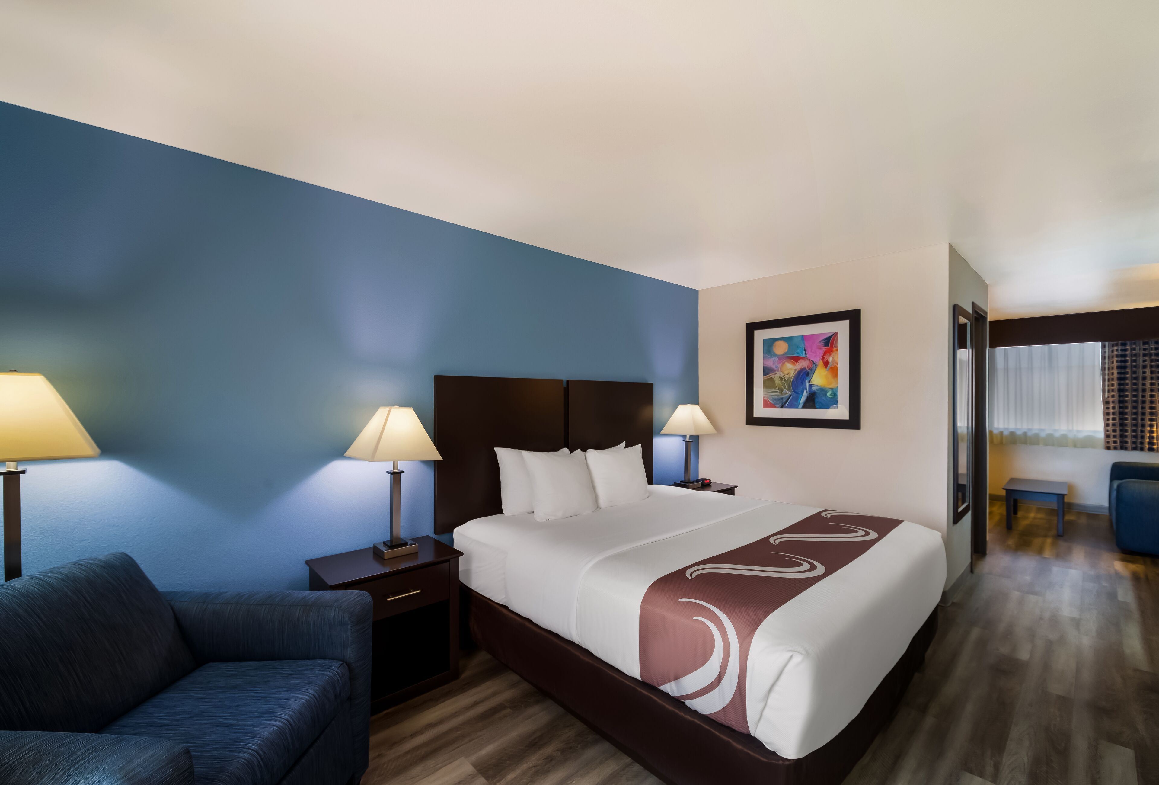 A hotel room with a large bed, two bedside tables with lamps, a blue wall, a painting, and a seating area with a chair and table.