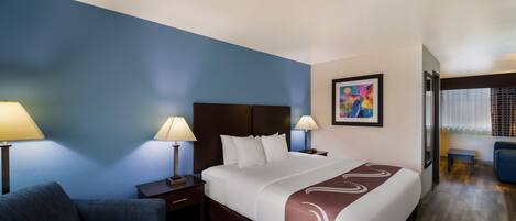A hotel room with a large bed, two bedside tables with lamps, a blue wall, a painting, and a seating area with a chair and table.