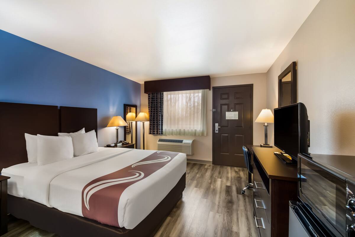 A hotel room with a large bed, a desk with a television, a desk lamp, and a window with curtains.