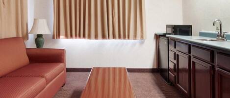 Habitación de hotel con sofá, mesa de madera, kitchenette y ventana con cortinas.