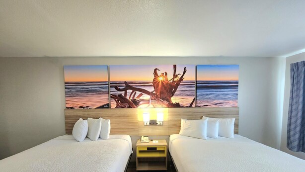 A hotel room with two beds, a wooden headboard, a nightstand with a lamp, and a large beach sunset painting on the wall. - Days Inn & Suites by Wyndham Corpus Christi Central (Corpus Christi)