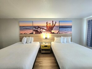 Habitación de hotel con dos camas, cabecera de madera, mesita de noche con lámpara y un cuadro grande de una puesta de sol en la playa.