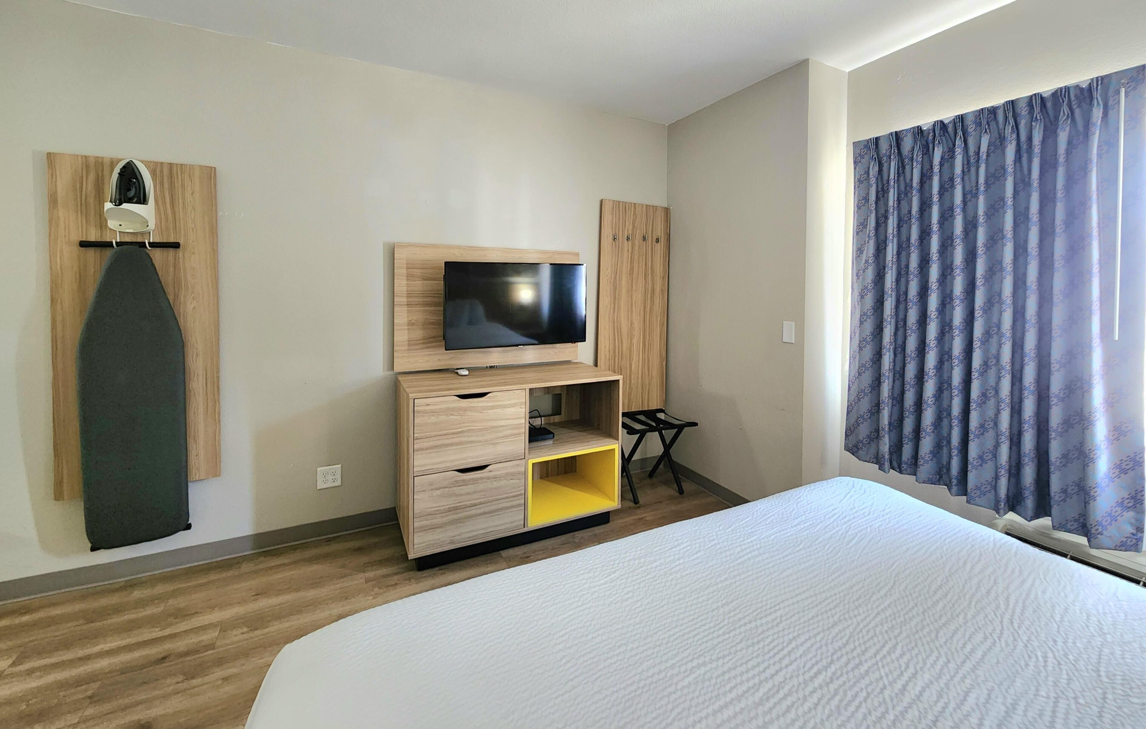A hotel room with a bed, a TV, a wooden wardrobe, a shelf, and a curtain.