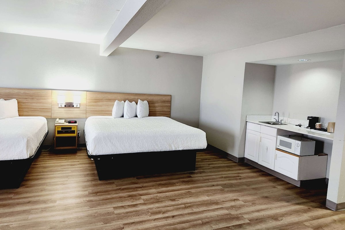 A hotel room with two beds, a wooden headboard, a small bedside table with a lamp, a built-in desk with a sink, and a microwave.