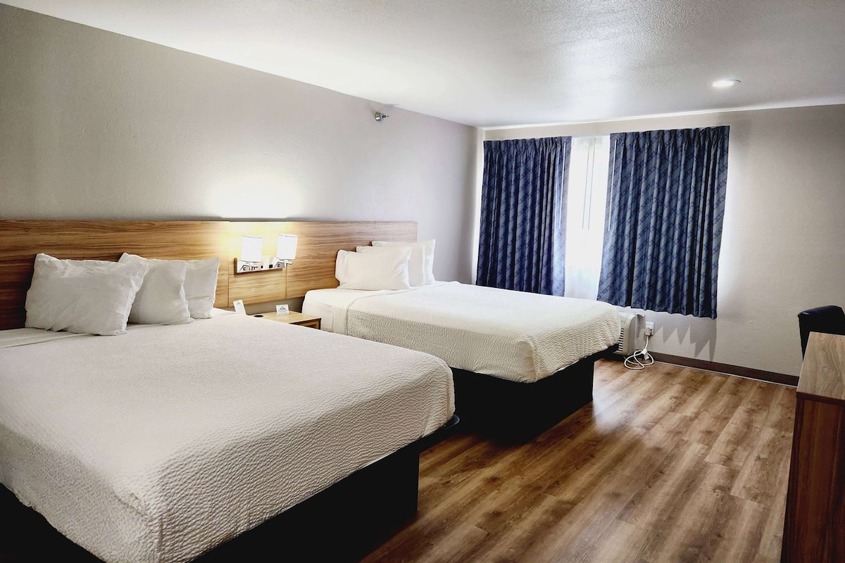 A hotel room with two beds, wooden headboards, white bedding, a nightstand, a lamp, and a window with curtains.