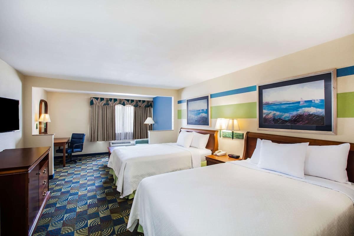 A hotel room with two beds, a desk, a chair, a TV, and artwork on the walls.