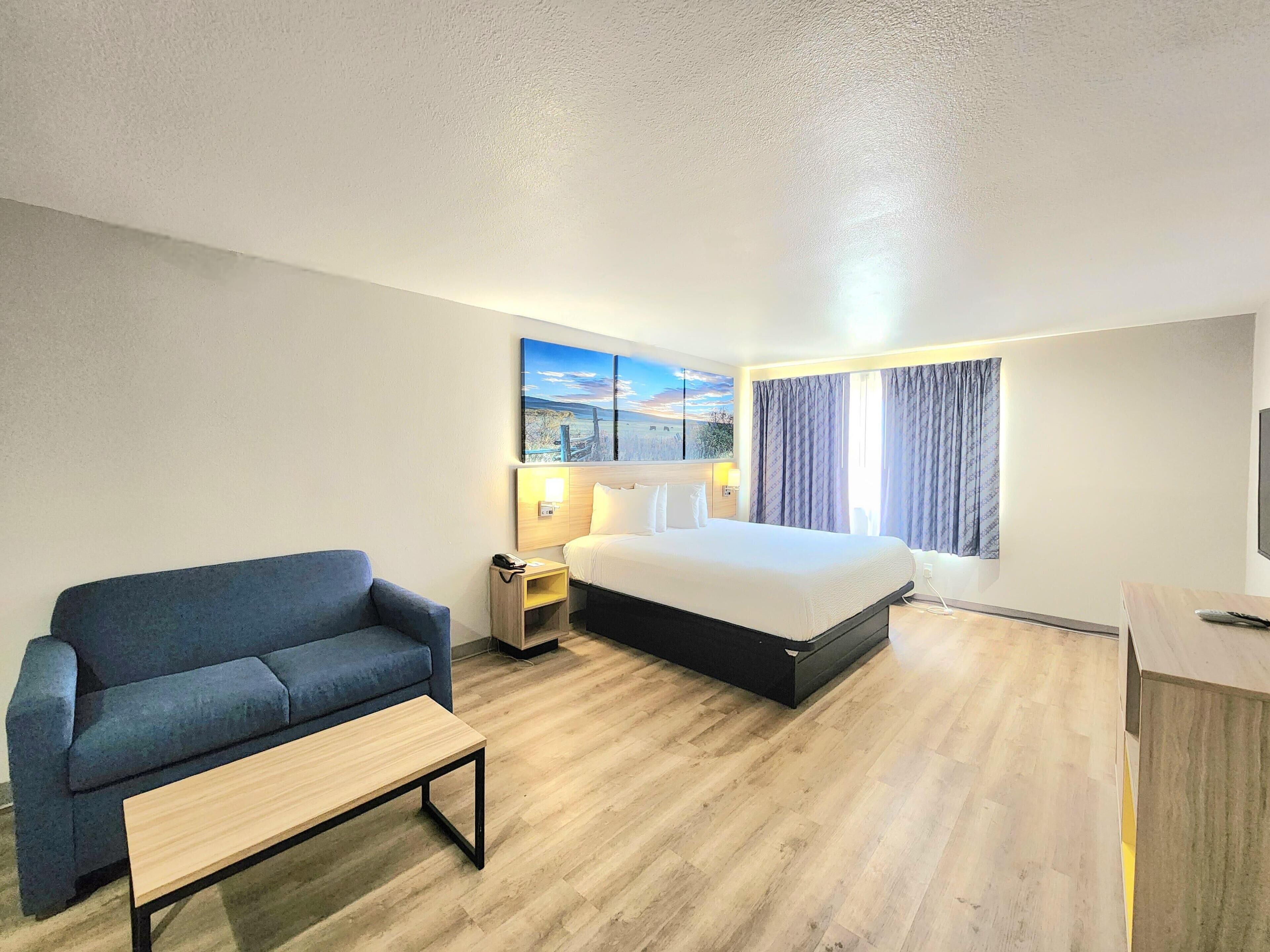 A hotel room with a bed, a sofa, a bedside table, a lamp, a painting, and a TV.
