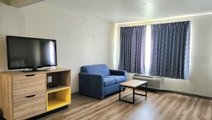 A living room with a television on a wooden stand, a blue sofa, a small table, and a window with curtains. - Days Inn & Suites by Wyndham Corpus Christi Central (Corpus Christi)