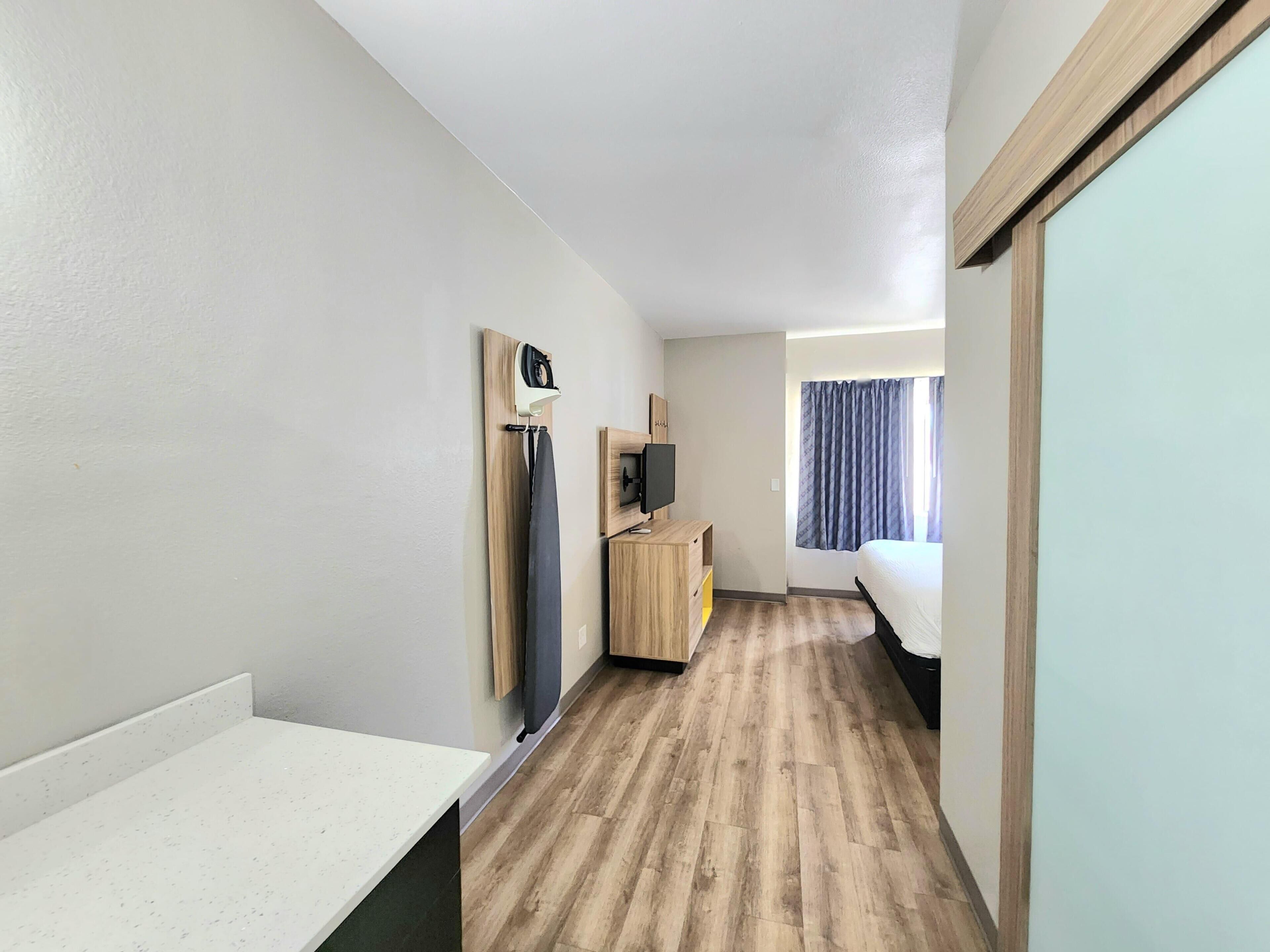 Una habitación de hotel estrecha con suelo de madera, una cama, un escritorio, un televisor y una ventana con cortinas.
