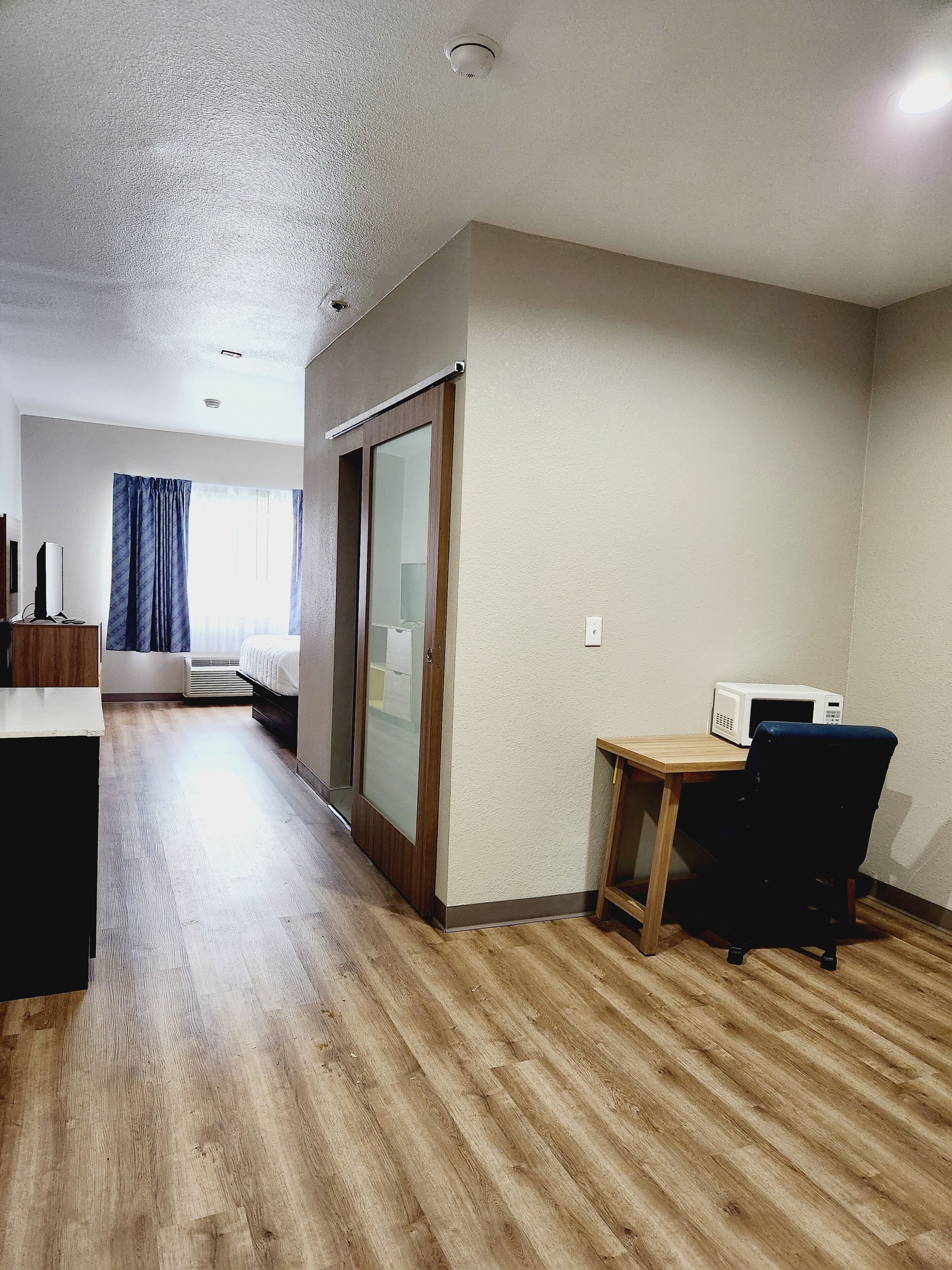 A hotel room with a wooden floor, a desk with a chair, a microwave, a sliding door, and a window with curtains.