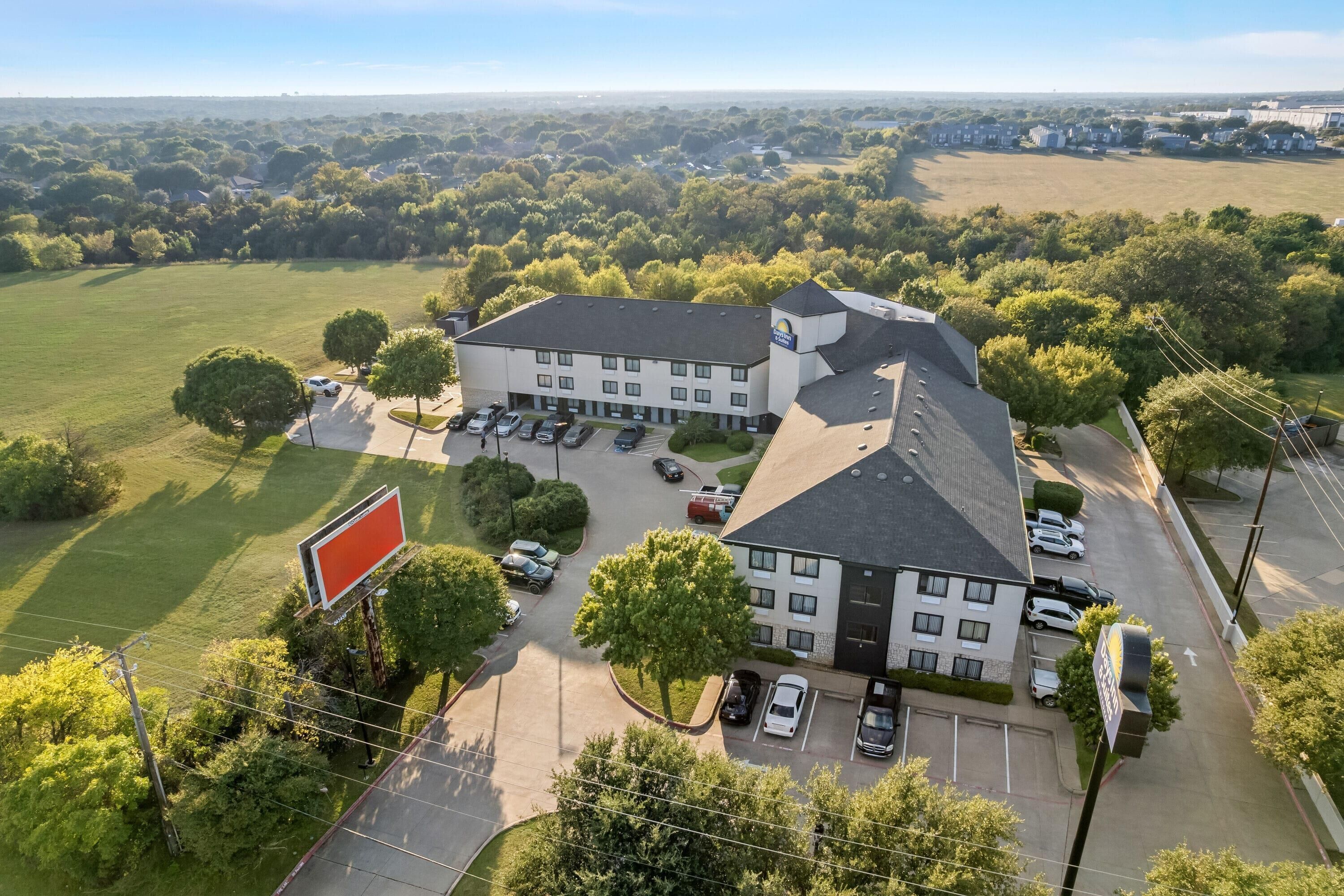 An aerial view of a hotel with a parking lot, surrounded by greenery and a clear sky.