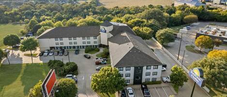 An aerial view of a hotel with a parking lot, surrounded by greenery and other buildings.