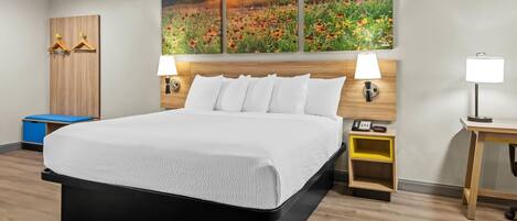 A hotel room with a large bed, two bedside lamps, a wooden headboard, a wall-mounted painting, and a small wooden nightstand with a lamp.