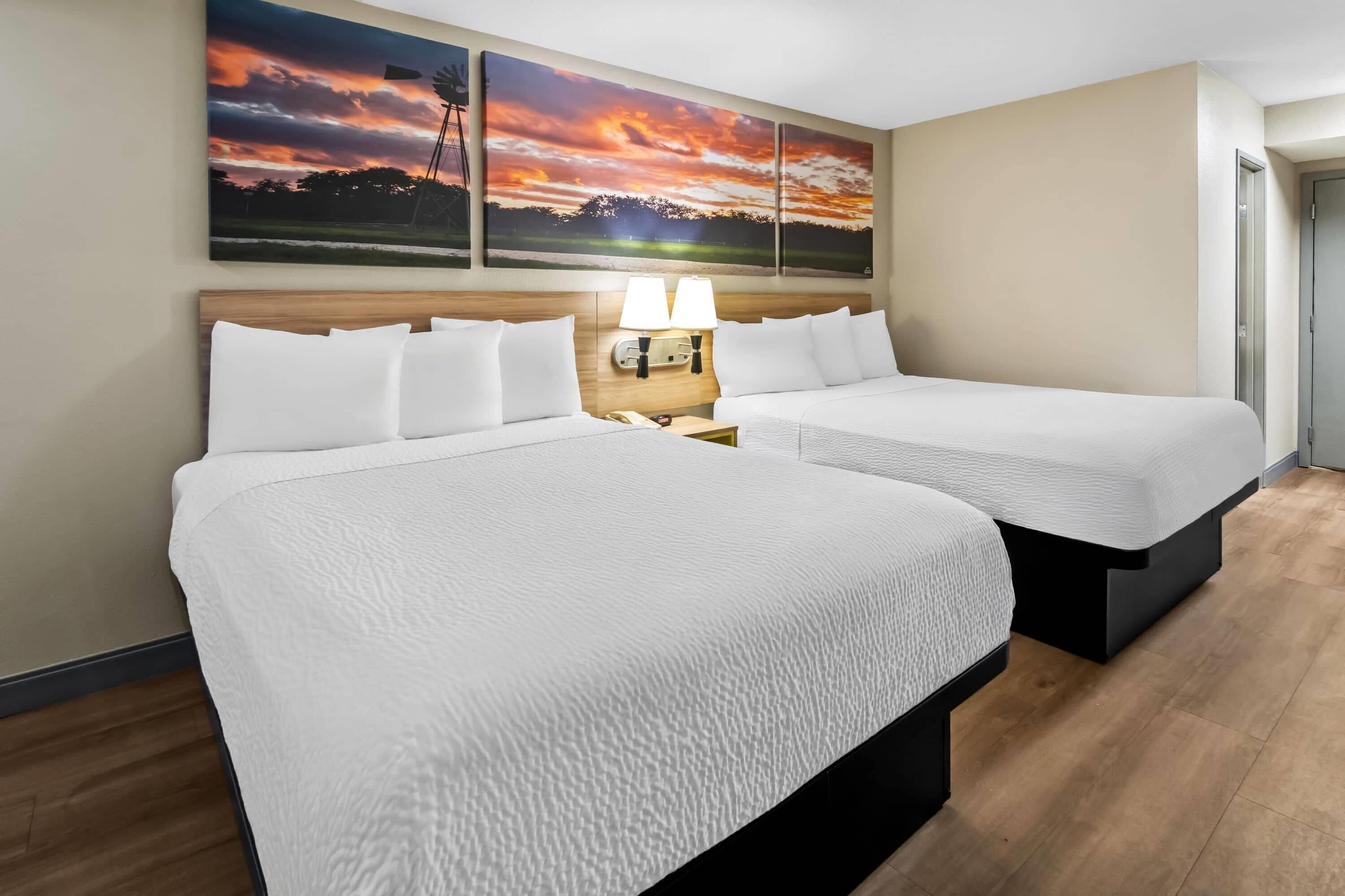 Ein Hotelzimmer mit zwei Betten, einem Nachttisch mit Lampe und einem Sonnenuntergangsbild an der Wand.