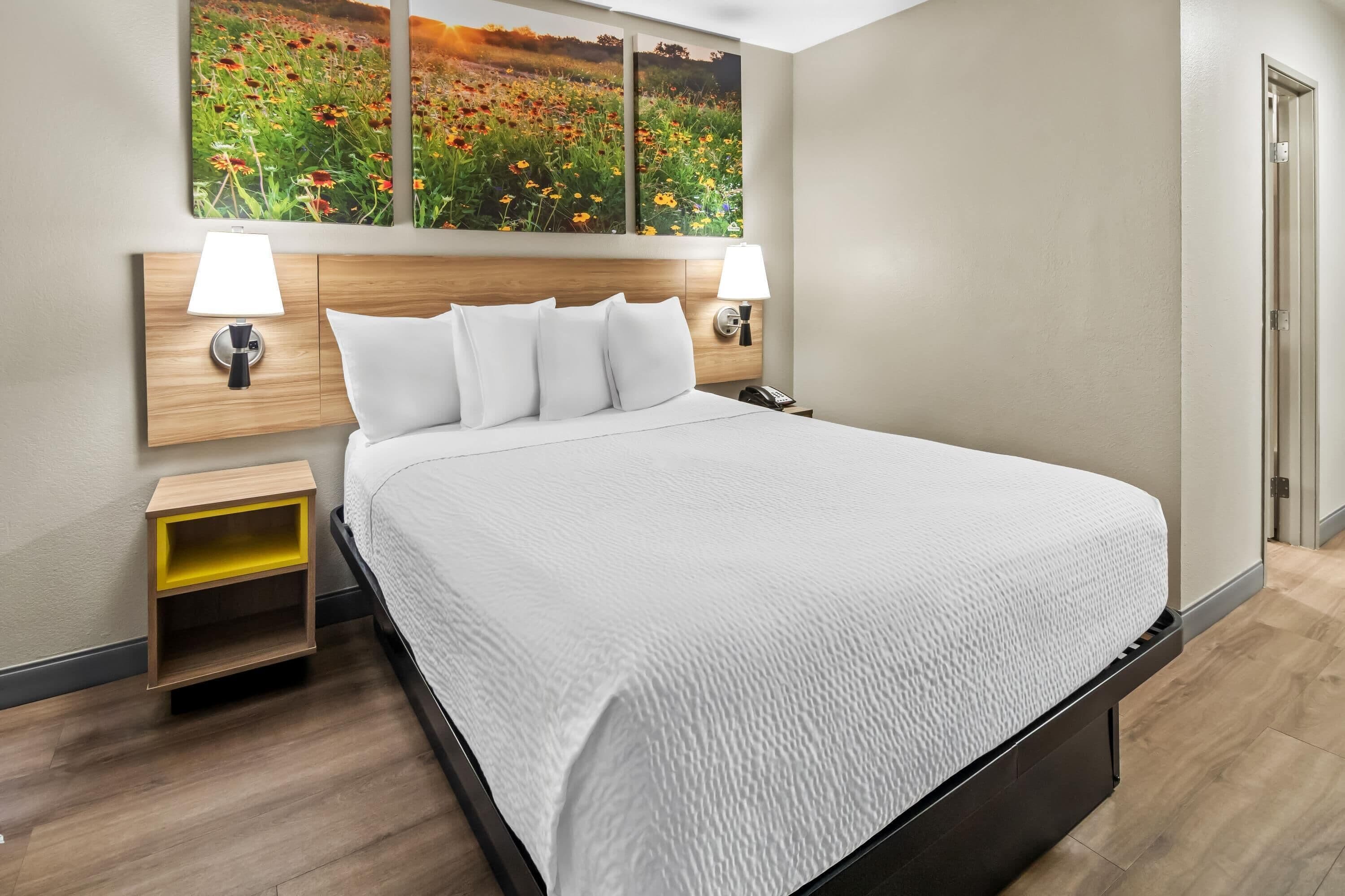 Habitación de hotel con una cama grande, dos mesitas de noche con lámparas, un cabecero de madera y una obra de arte en la pared que representa un campo.