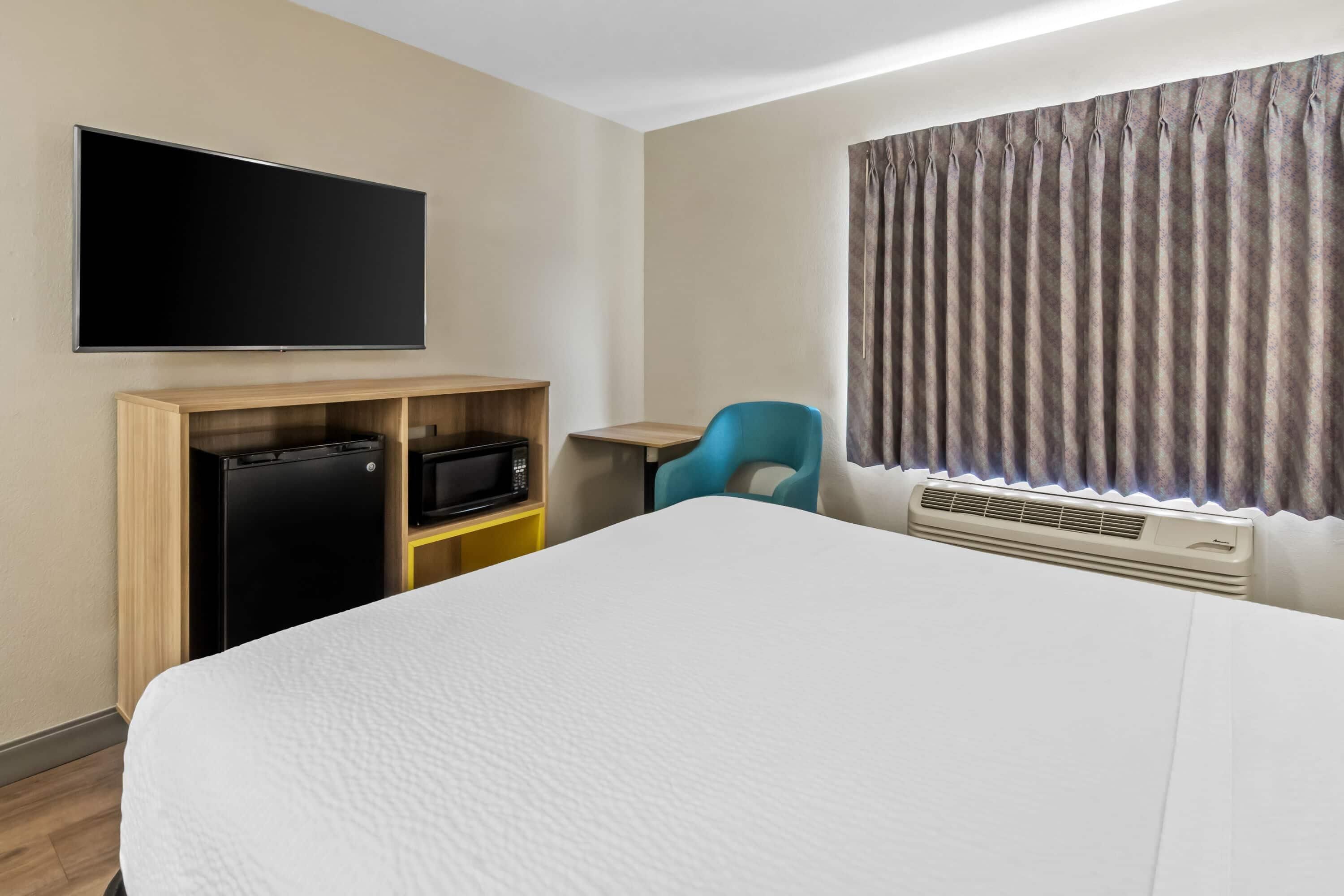 A hotel room with a bed, a flat-screen TV mounted on the wall, a small desk with a chair, a microwave, and a mini-fridge.