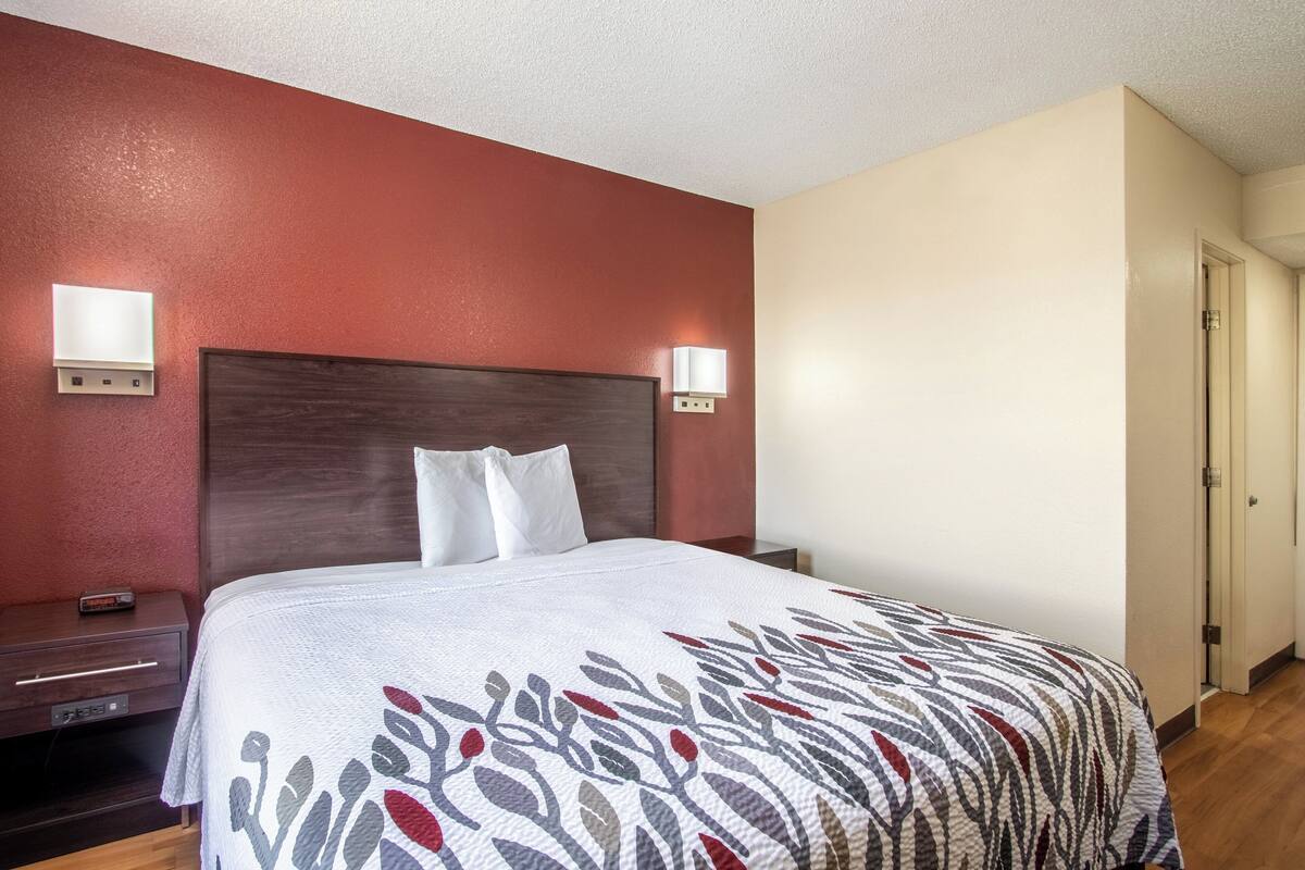 A hotel room with a large bed, a nightstand, a wall-mounted lamp, and a door.