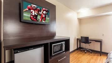 A modern hotel room with a flat-screen TV mounted on a wooden cabinet, a built-in mini-fridge, a microwave, and a desk with a chair.