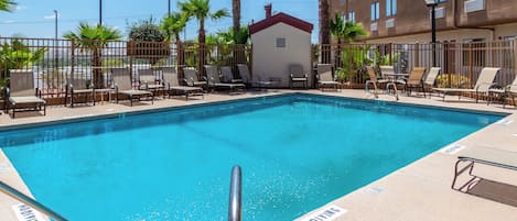 A rectangular swimming pool with a metal ladder, surrounded by lounge chairs and palm trees, with a building in the background.