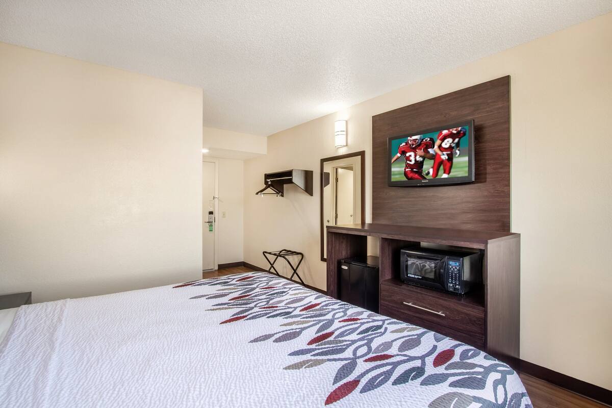 A hotel room with a bed, a flat-screen TV mounted on the wall, a microwave, and a small seating area with a chair.