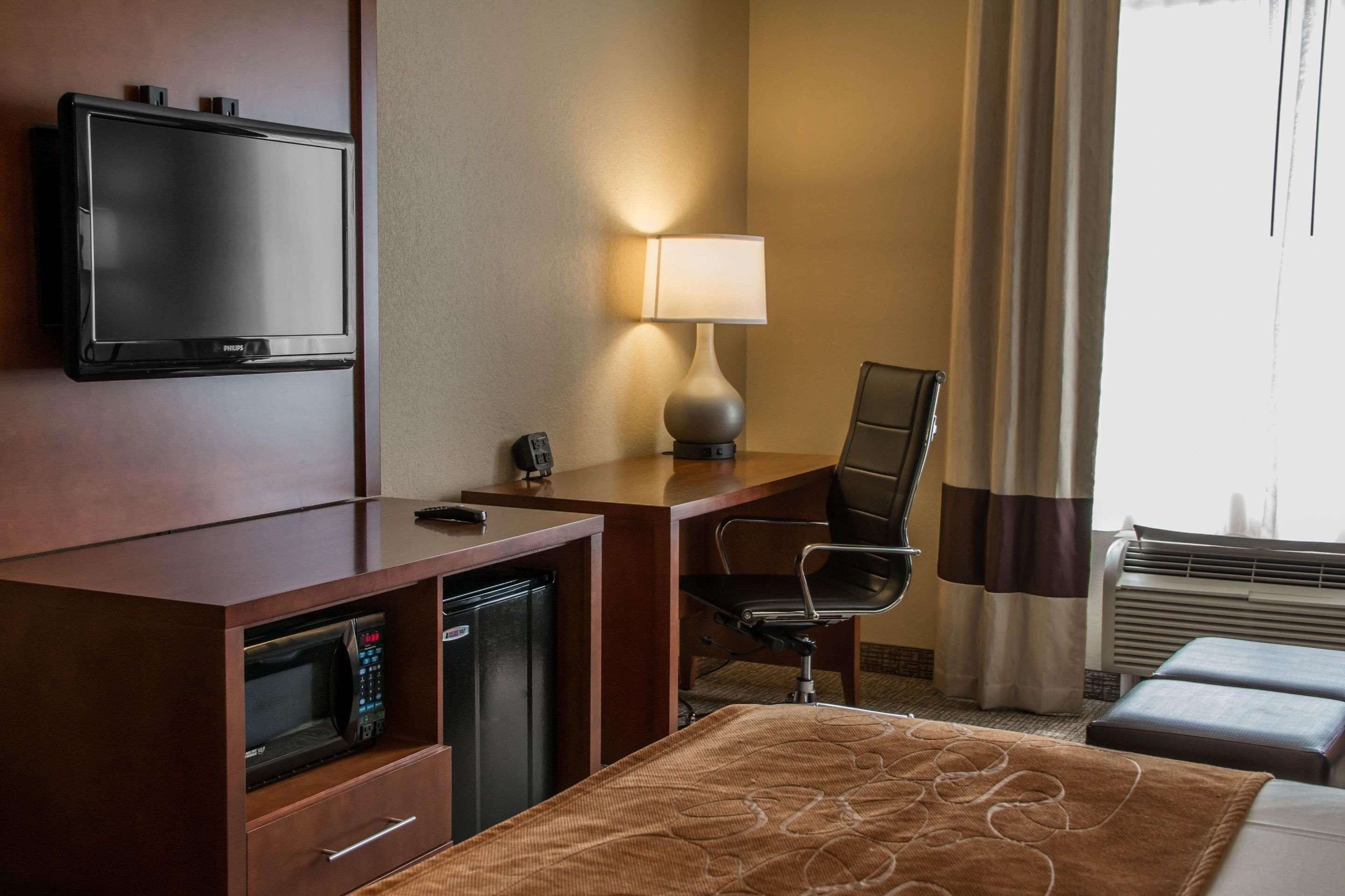 Quarto de hotel com televisão de tela plana, escrivaninha com luminária, micro-ondas, mini-geladeira, cadeira, cama com manta marrom e janela com cortinas.