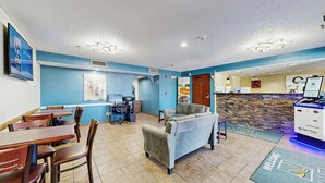 A hotel lobby with a reception desk, a seating area with a sofa and chairs, a dining area with tables and chairs, and a stone wall with a welcome mat.