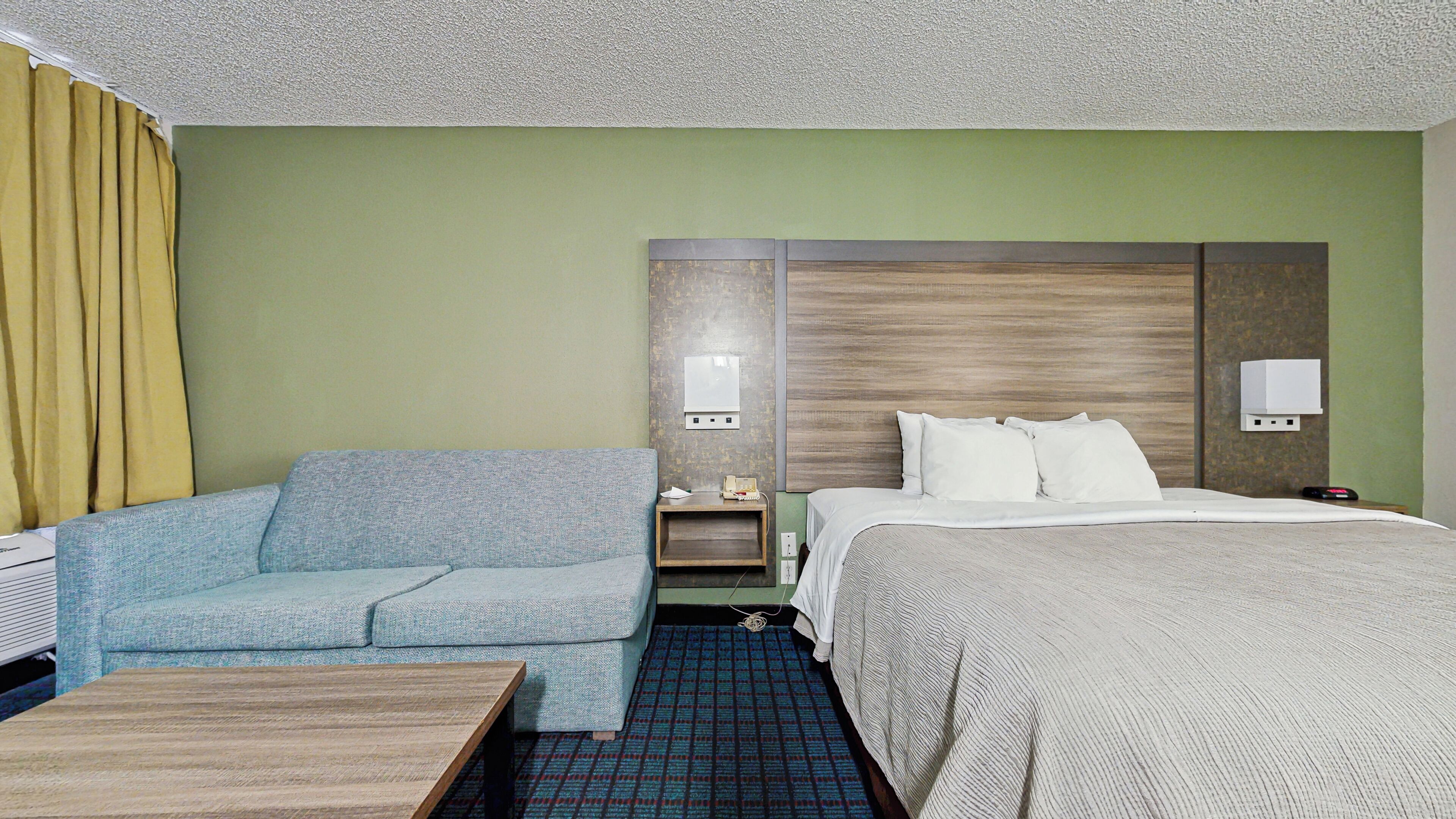 A hotel room with a bed, a sofa, a small table, and a nightstand with a lamp.
