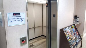 An elevator interior with a sign instructing to use the stairs in case of fire, a fire alarm button, and a magazine rack with various publications. - Coratel Inn & Suites by Jasper New Braunfels IH-35 EXT 189 (New Braunfels)