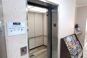 An elevator interior with a sign instructing to use the stairs in case of fire, a fire alarm button, and a magazine rack with various publications.