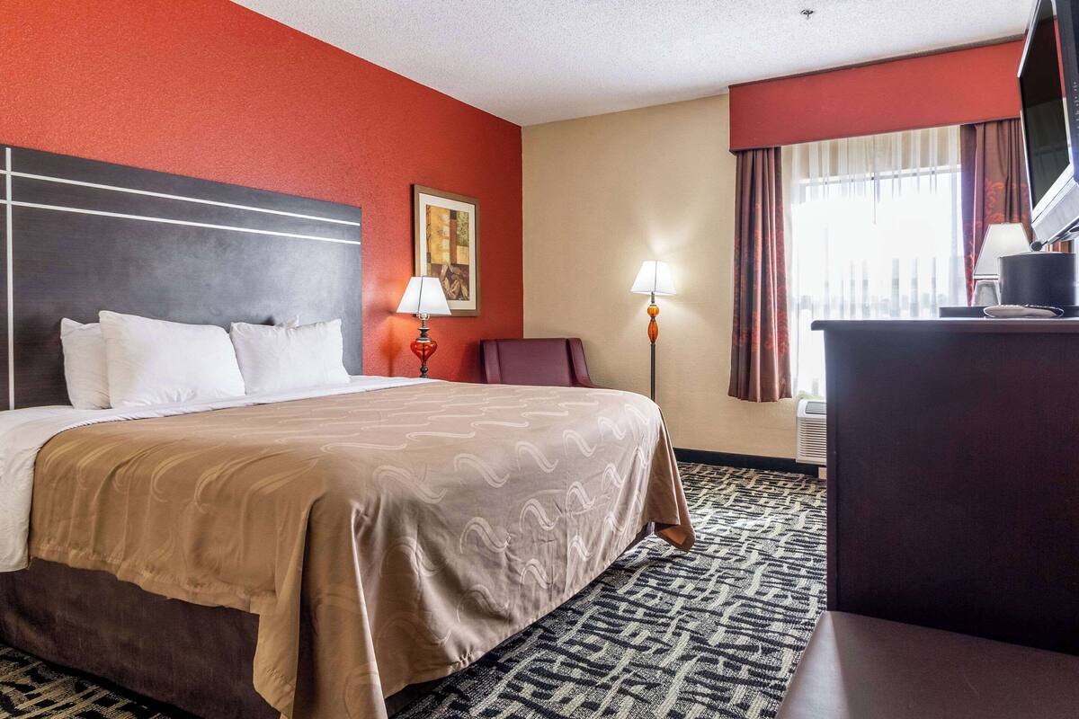 A hotel room with a large bed, a desk, a chair, a TV, and a window with curtains.