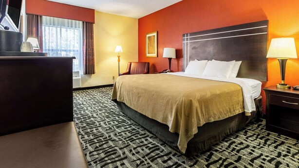 A hotel room with a large bed, a desk, two bedside lamps, a window with curtains, and a patterned carpet.