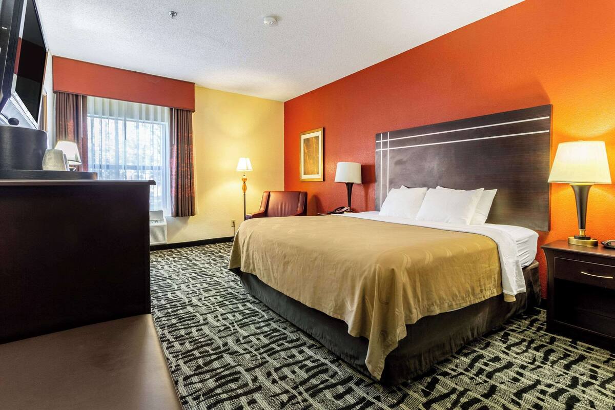 A hotel room with a large bed, a desk, two bedside lamps, a window with curtains, and a patterned carpet.