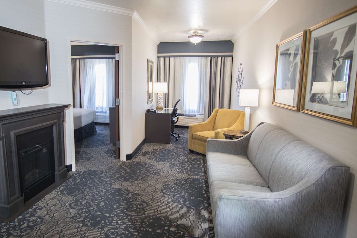 A hotel room with a flat-screen TV, a fireplace, a sofa, a yellow armchair, a desk with a lamp, and a large mirror.