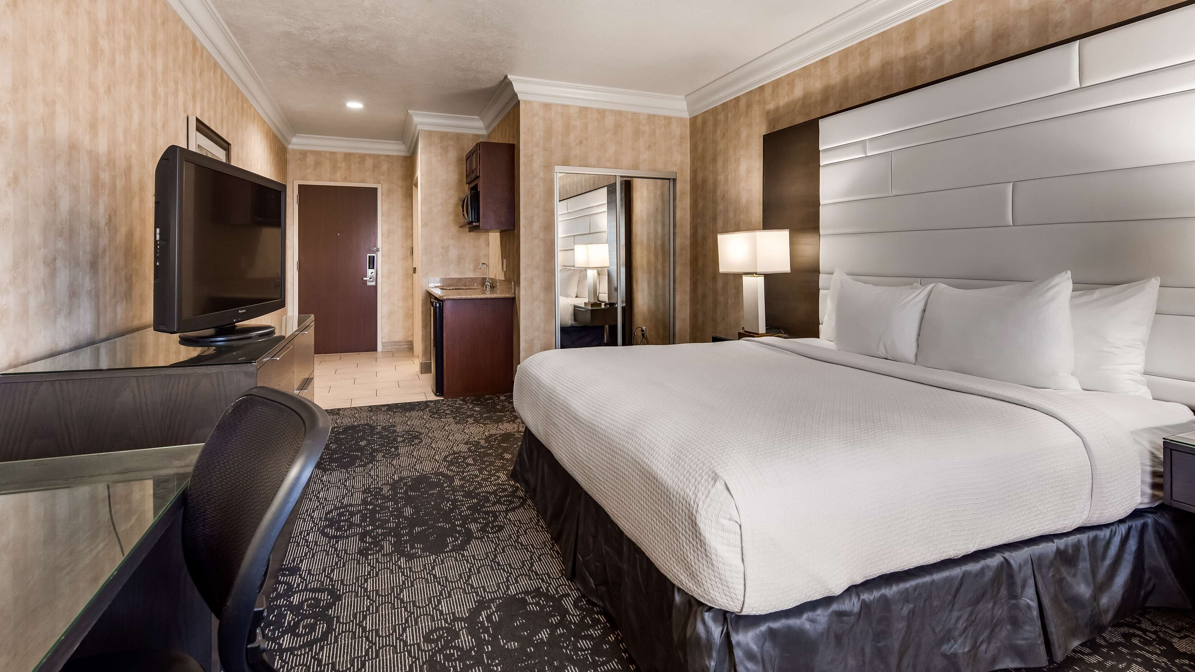 Une chambre d’hôtel avec un grand lit, un bureau doté d’un téléviseur à écran plat, un miroir, une table de chevet avec une lampe, et une salle de bain visible à travers une porte ouverte.