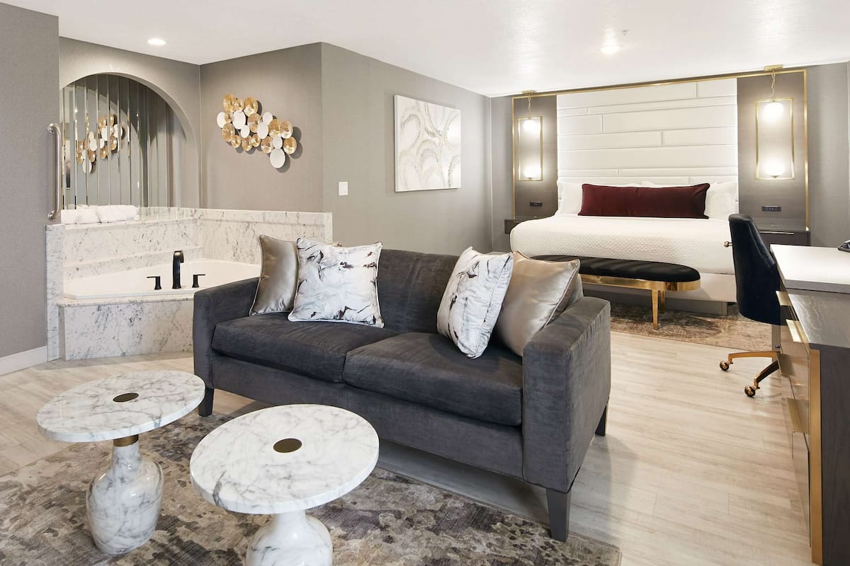 A modern hotel room with a grey sofa, two marble coffee tables, a bed with a white headboard, a desk with a chair, and a bathroom with a marble sink and vanity.