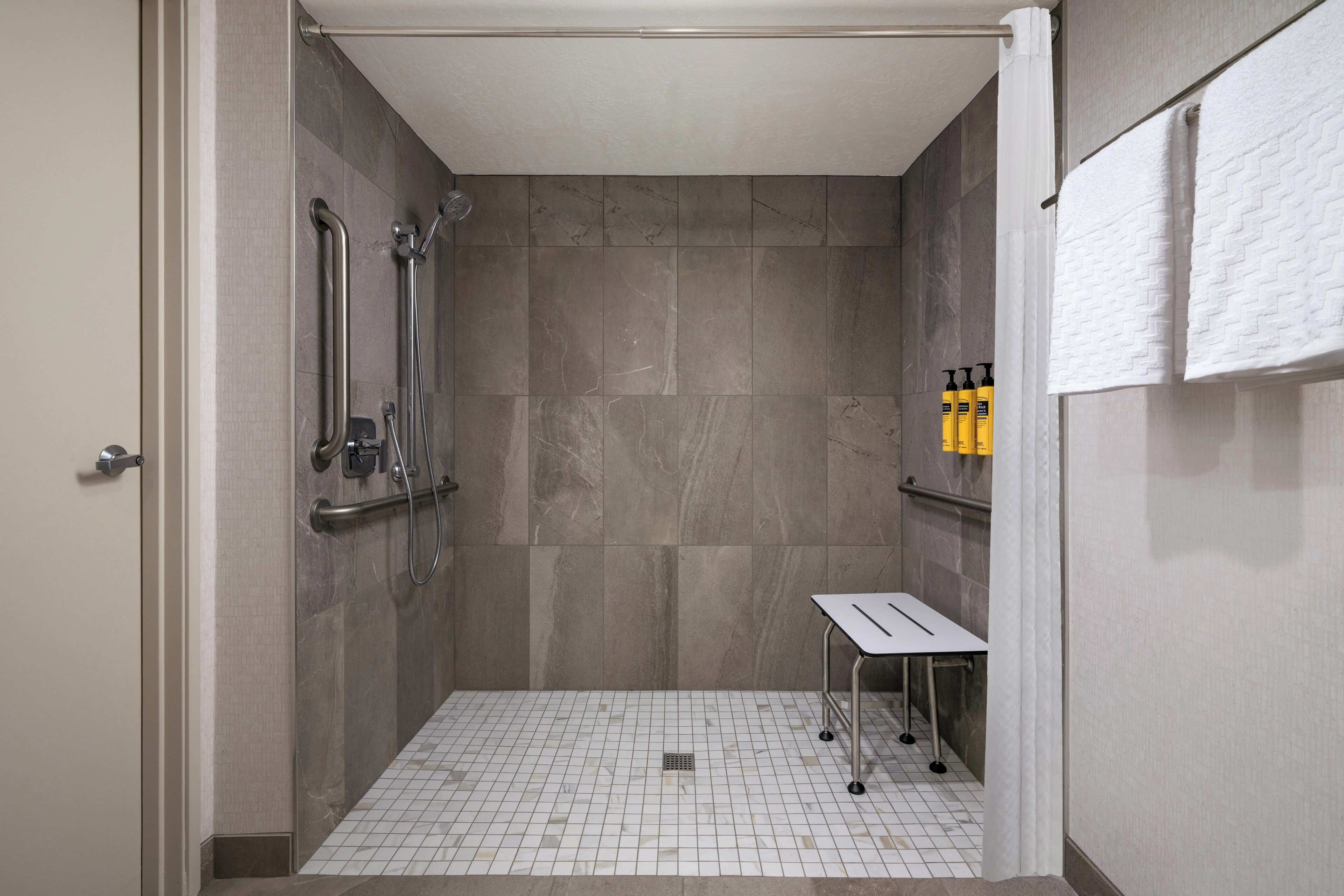 A shower with a handrail, a white tiled floor, and a small table with a towel rack.