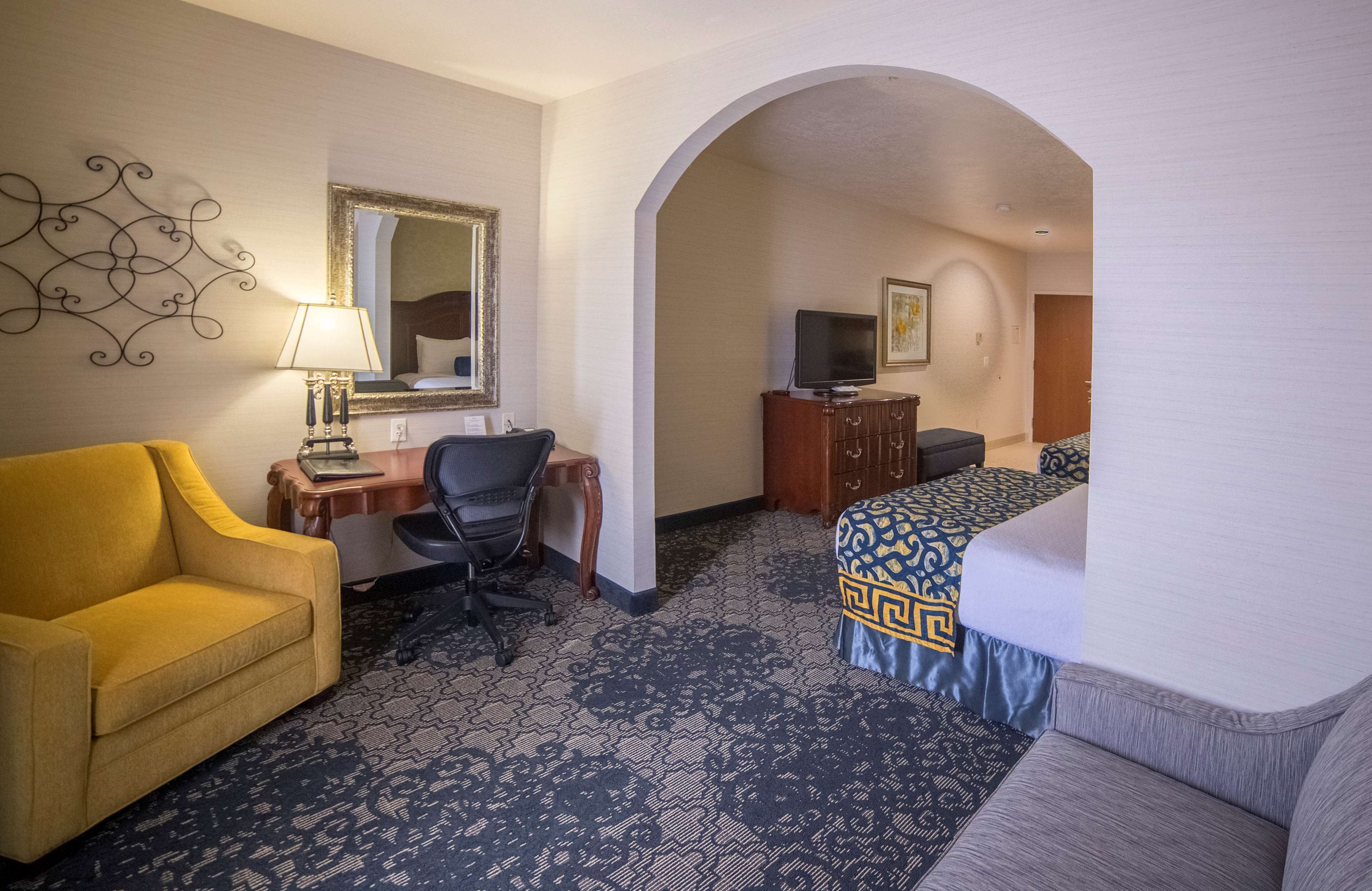 A hotel room with a yellow armchair, a desk with a lamp, a computer, a TV, a bed with a patterned bedspread, and a patterned carpet.