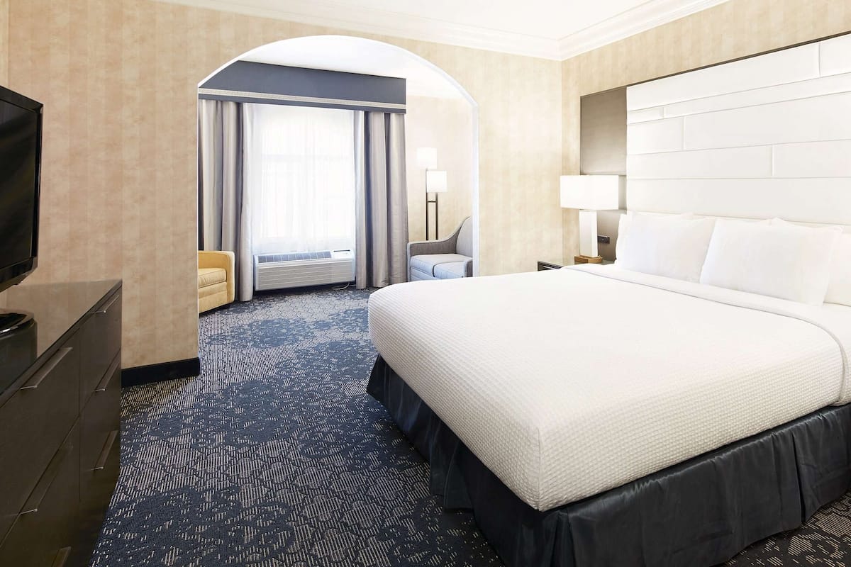 A hotel room with a large bed, a TV, a desk with a lamp, and a view through an arched doorway.