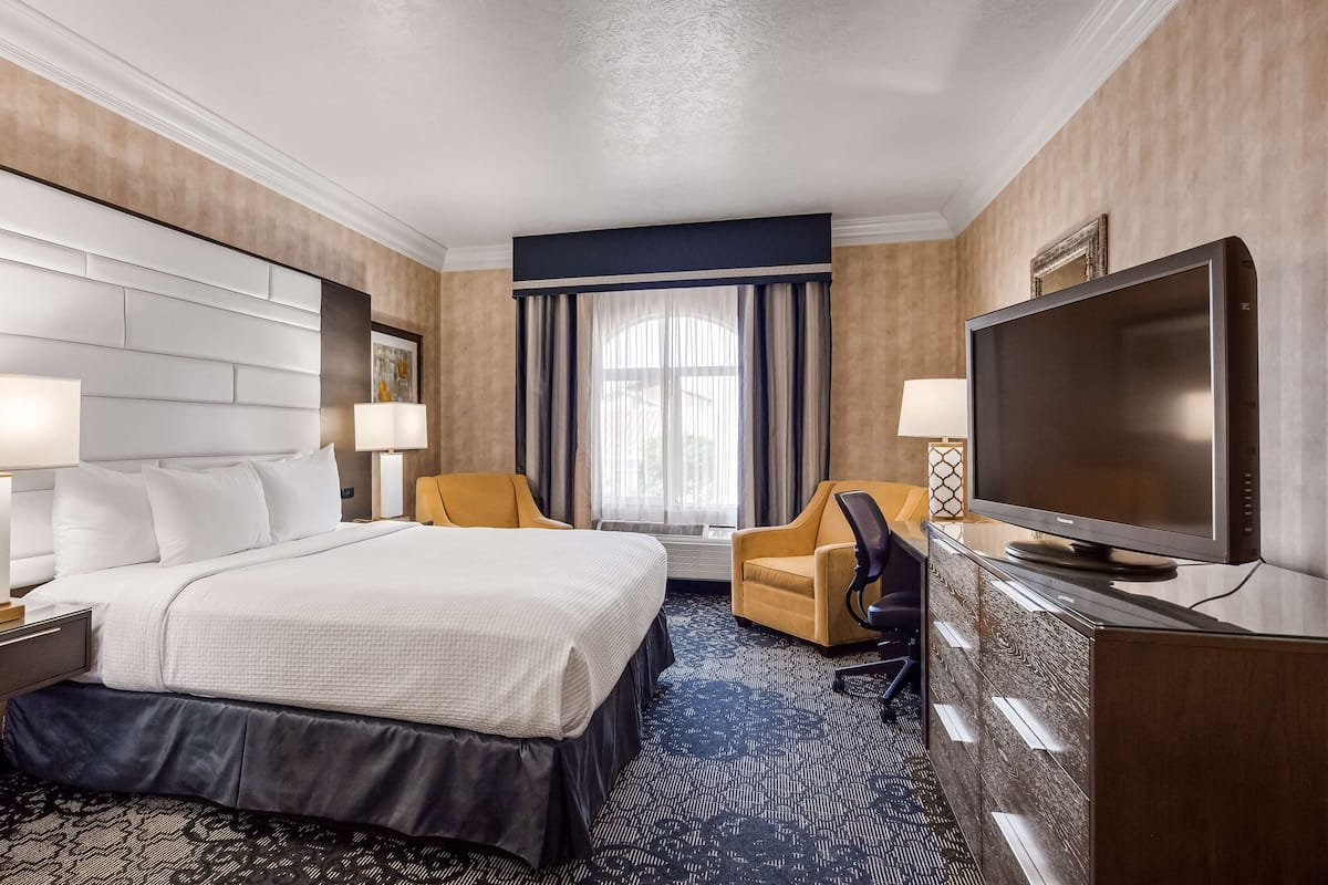 A hotel room with a large bed, two chairs, a desk with a flat-screen TV, and a large window with curtains.