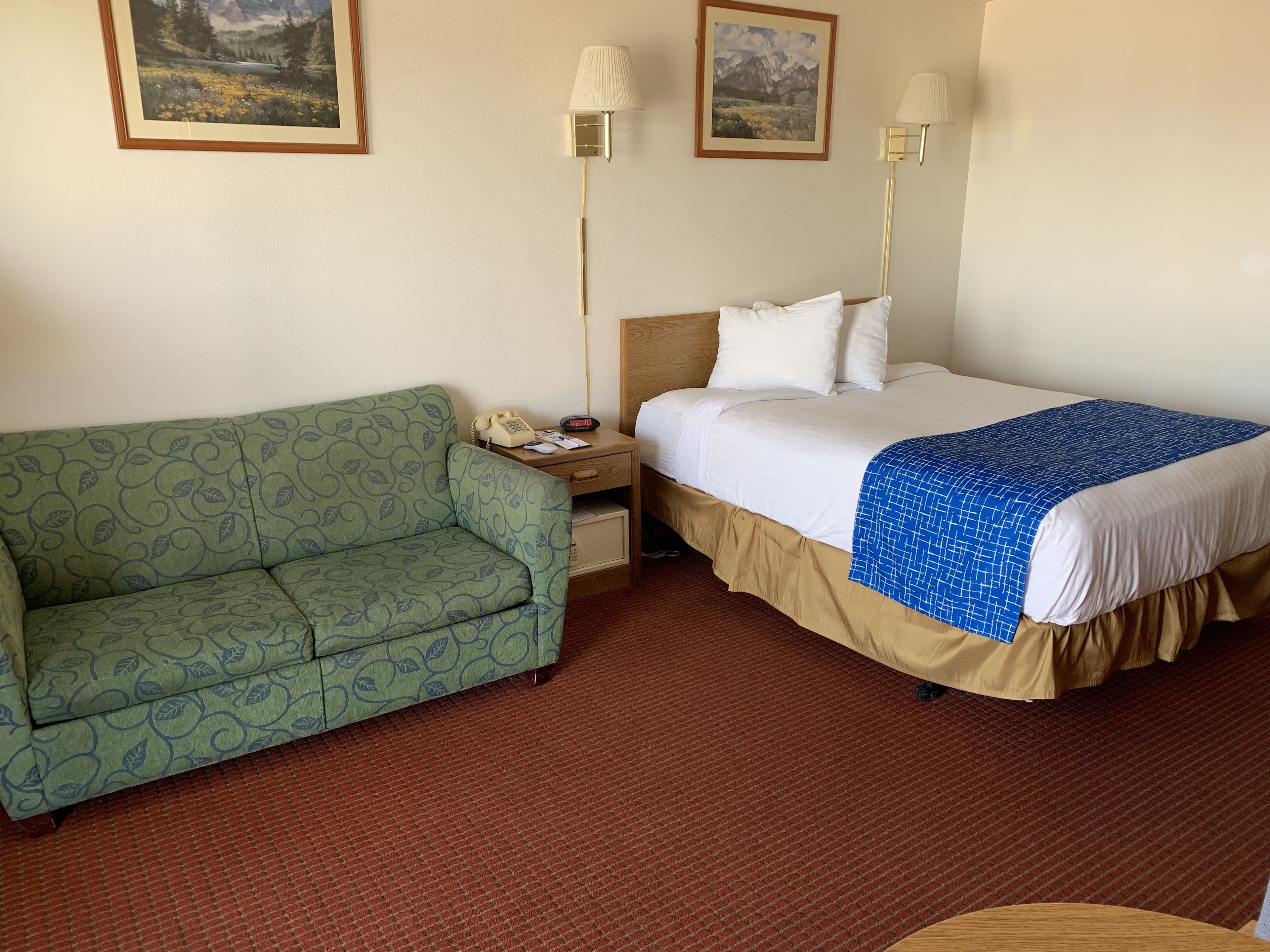 A hotel room with a green sofa, a bed with a blue and white patterned bedspread, two wall-mounted lamps, and two framed landscape paintings.