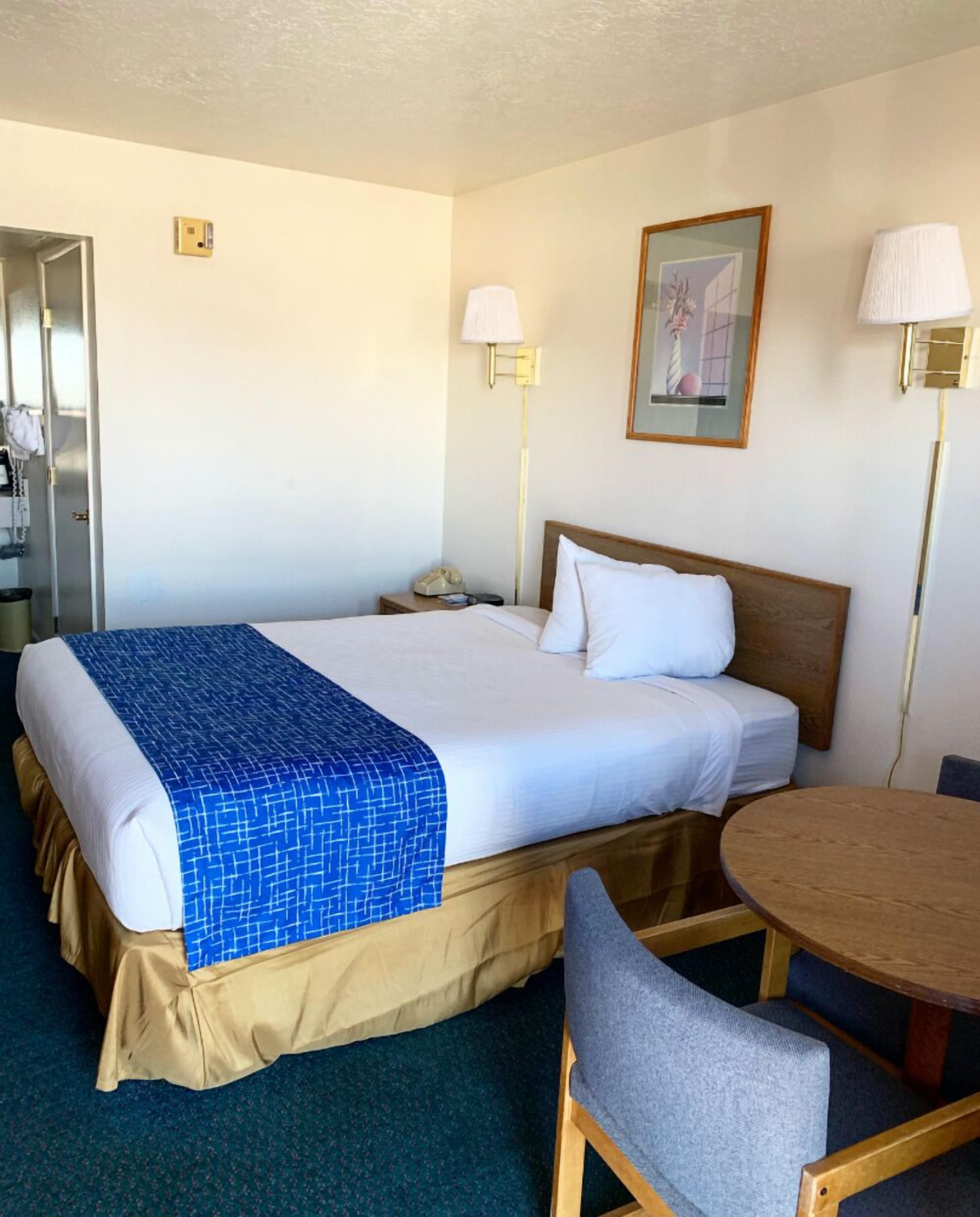 Una habitación de hotel con cama, mesita de noche, lámpara, silla y una pequeña mesa redonda.