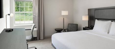 A hotel room with a large bed, a desk, a chair, a lamp, and a view of greenery through the window.