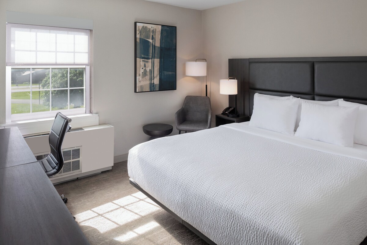 A hotel room with a large bed, a desk, a chair, a nightstand with a lamp, and a window with blinds.
